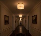 Cream alabaster flush mount light repeats along a dim hallway with framed paintings, paneled walls, and dark wooden flooring.