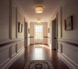 Cream alabaster flush mount light hangs in a sunlit hallway with white paneled walls, framed pictures, and wooden flooring.