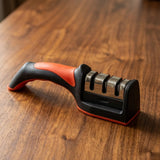 A handheld sharpening stone set with a black and orange ergonomic handle, resting on a wooden tabletop.