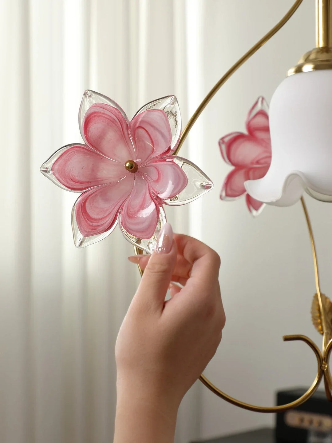 A hand touches a pink glass flower on a gold pendant light beside its white scalloped shade against pale curtains.