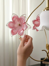 A hand touches a pink glass flower on a gold pendant light beside its white scalloped shade against pale curtains.