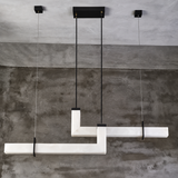 Modern geometric linear pendant light fixture with white marble-like horizontal bars suspended by black cables against a textured concrete wall and ceiling.