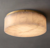 Low-angle view of an illuminated cream alabaster flush mount light with a gold-colored rim and broad circular underside against gray.