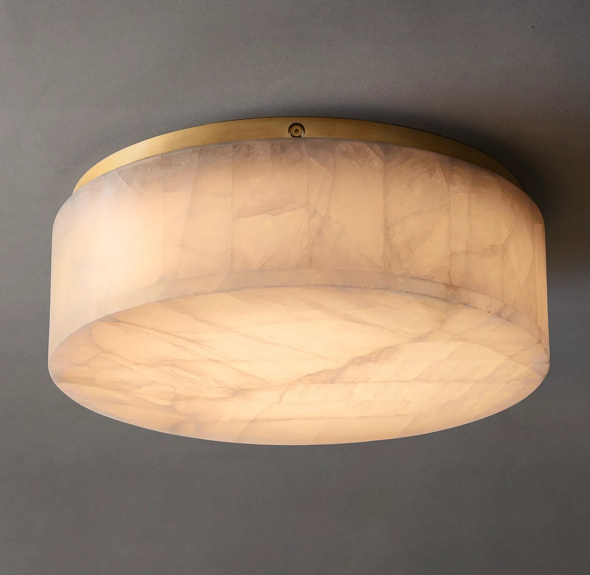 Low-angle view of an illuminated cream alabaster flush mount light with a gold-colored rim and broad circular underside against gray.