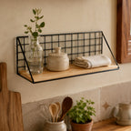 Black wire grid wall shelf with wood base holding a glass bottle, a stoneware pot and a folded cloth