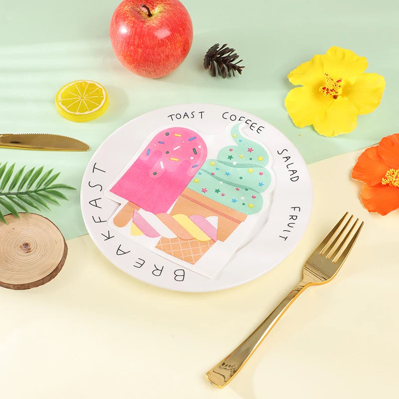A white paper plate with a colorful ice cream illustration and the words 'TOAST COFFEE SALAD FRUIT BREAKFAST' arranged on a light cream surface, surrounded by a gold fork, red apple, yellow flower, orange flower, lemon slice, pinecone, and wood slice on a pale mint background.