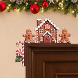 Brown gingerbread house door corner decoration with two smiling figures and a hanging candy cane sits on a wooden doorframe beneath garland.