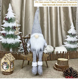 Gray gnome with dangling legs sits on a rustic chair among Christmas decorations, with a size label reading 16 × 46.5 cm.