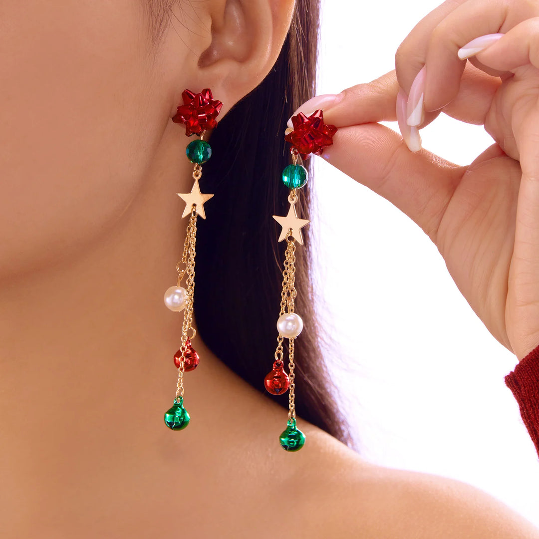 A person wears one red bow drop earring and holds its match, showing gold-tone stars, white beads, and red and green bells.