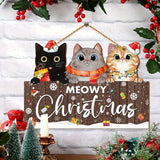Brown Meowy Christmas hanging sign with three cats hangs against white brick, surrounded by red berries, miniature trees, and a candle.