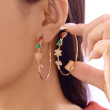 A model wears one gold-tone drop earring and holds another, both hoop-shaped with red bells, green trees, snowflakes and candy canes.