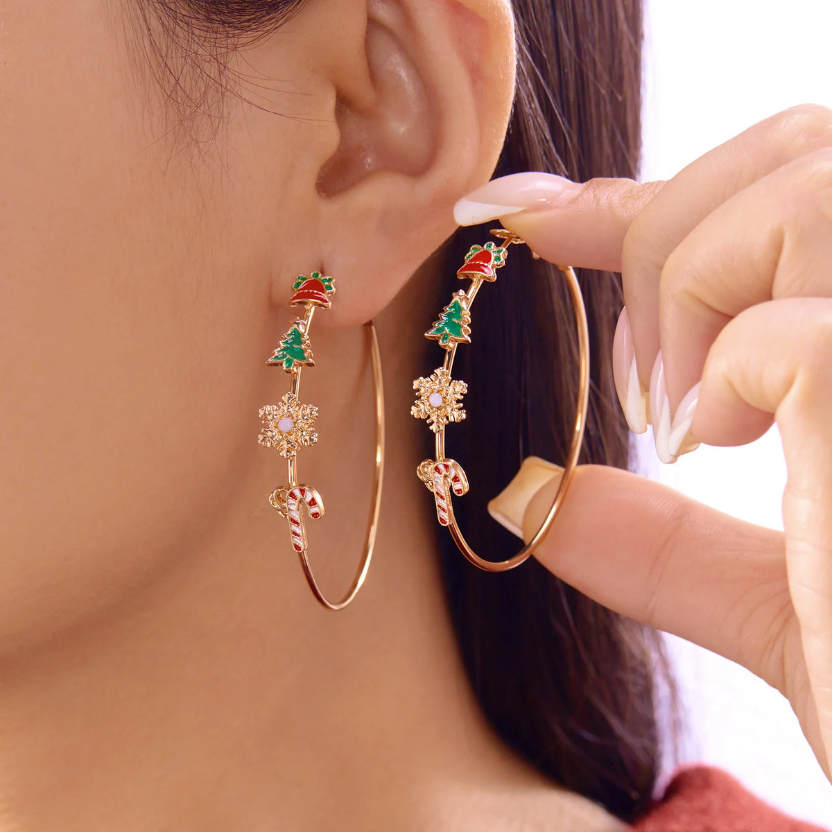 A model wears one gold-tone drop earring and holds another, both hoop-shaped with red bells, green trees, snowflakes and candy canes.