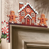 Gingerbread house corner decoration with white icing details and two smiling figures rests on beige door trim against a woodgrain wall.