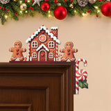Brown gingerbread house door corner decoration sits on dark wooden door trim beneath garland decorated with red and gold baubles.
