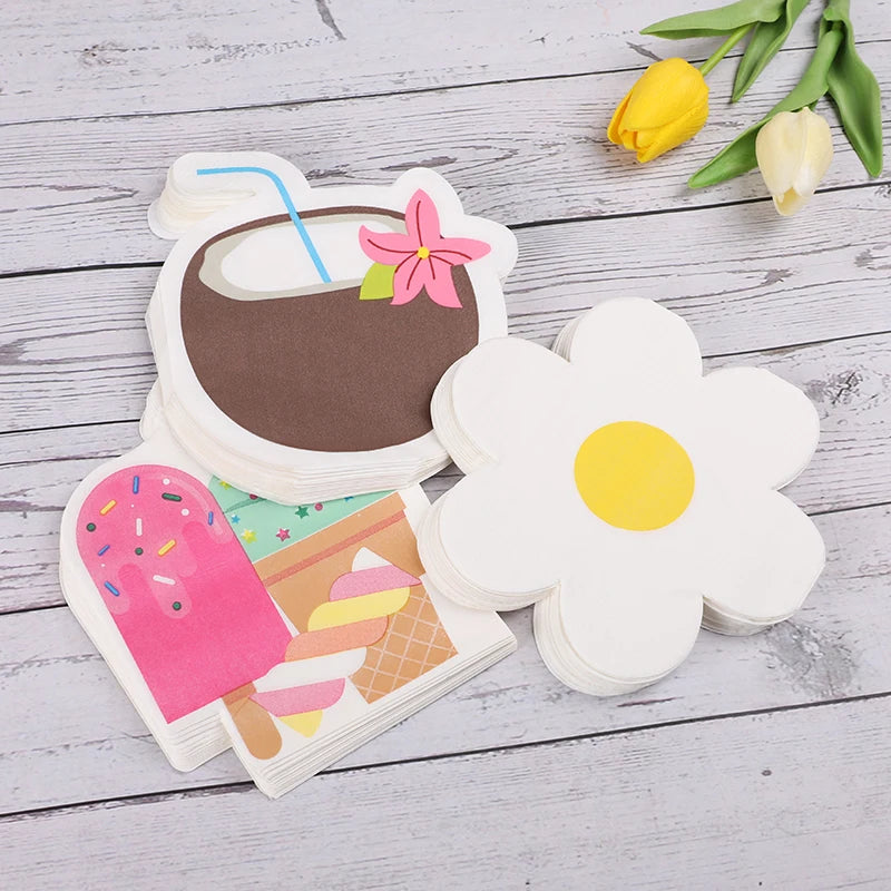 A set of cute, die-cut paper napkins featuring cartoon illustrations, including a coconut drink with a pink flower, a pink popsicle, and a white daisy flower. The napkins are arranged on a light gray wooden background with yellow tulips in the corner.