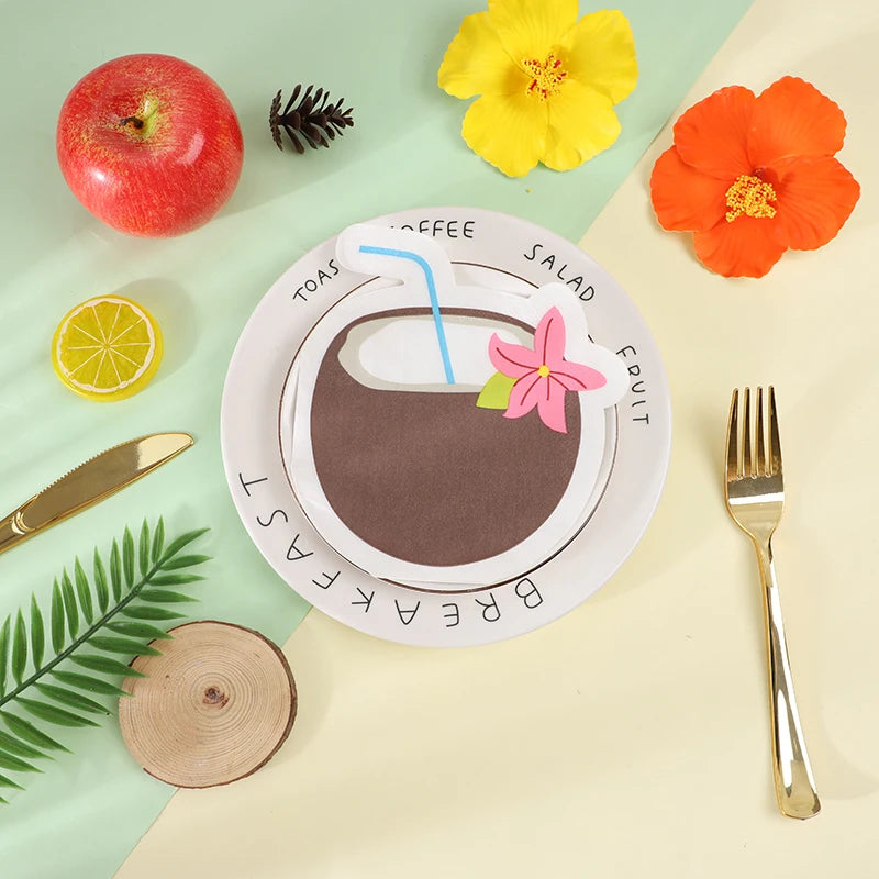A styled flat-lay of a white paper plate cutout shaped like a coffee cup with a straw, surrounded by tropical and breakfast-themed props including a red apple, yellow and orange flowers, a lemon slice, a knife, a fork, green leaves, and a wood slice on a split mint and cream background.
