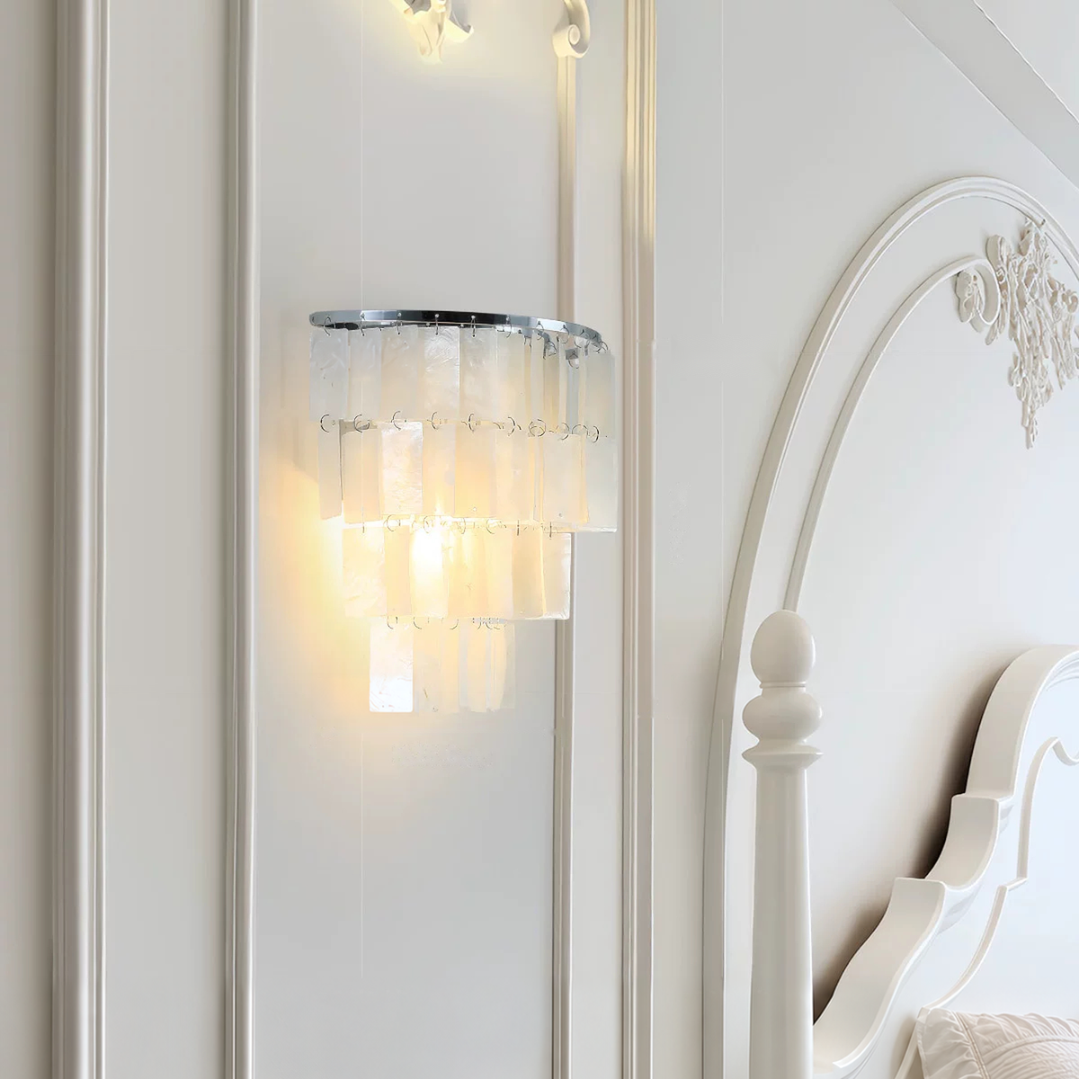 A modern wall sconce light fixture with a curved silver metal frame and multiple tiers of translucent white crystal or glass shades, glowing warmly against a white paneled wall next to a white bed.