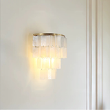 A modern wall sconce light fixture with a brushed gold metal frame and multiple tiers of translucent white textured glass prisms, shown illuminated on a white wall.