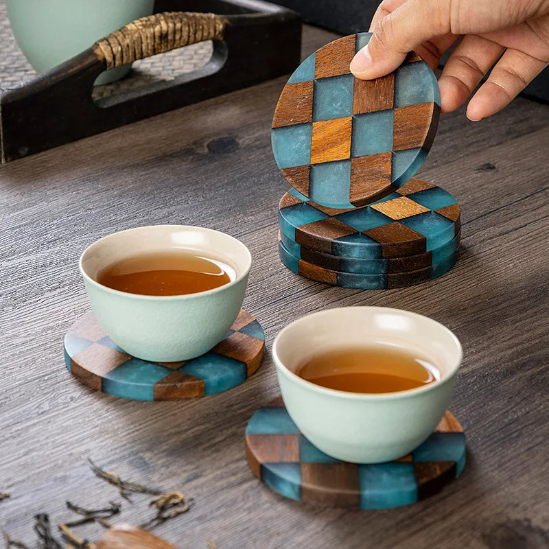 A hand holds a stack of round wooden coasters with a blue-green and brown checkerboard pattern on a dark wood table. Two pale green ceramic teacups filled with amber tea rest on matching coasters nearby.