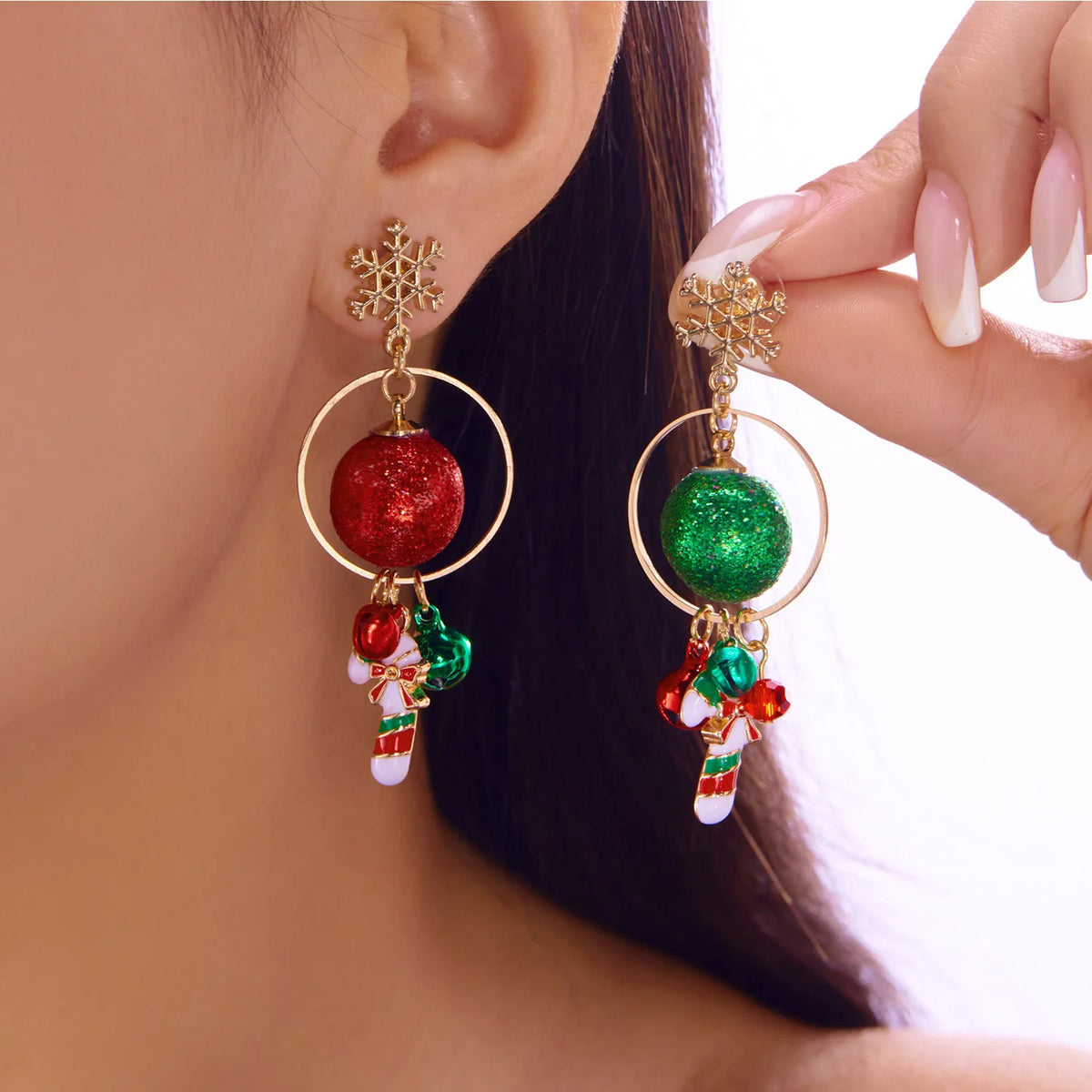 A model displays gold-tone drop earrings with snowflake studs, circular frames, red and green glitter baubles, bells, and striped candy cane charms.