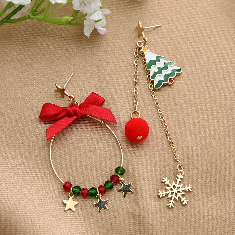 Gold-tone drop earrings with a red bow, beads, stars, Christmas tree, red bauble and snowflake rest on beige fabric beside white flowers.