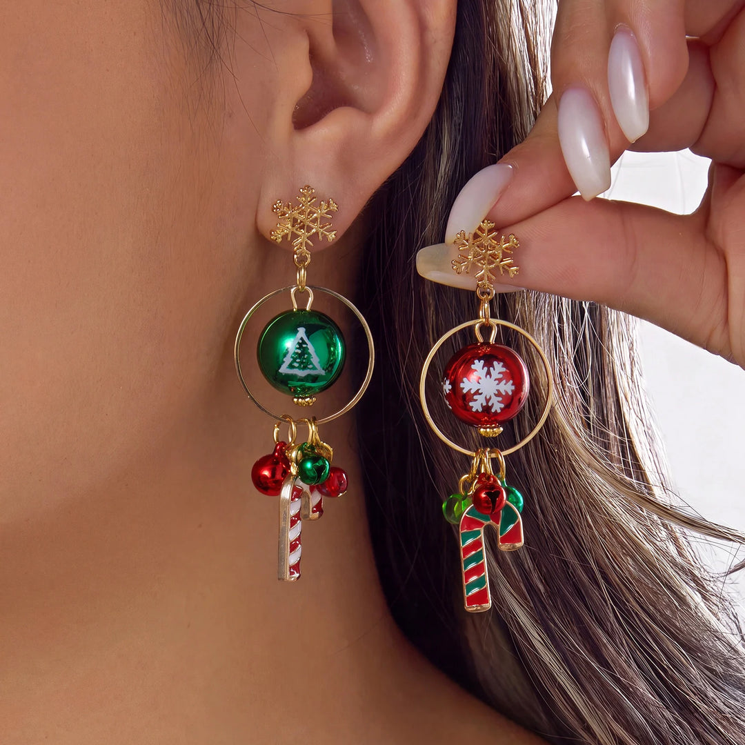 A model displays gold-tone drop earrings with snowflake studs, circular frames, printed red and green baubles, bells, and candy cane charms.