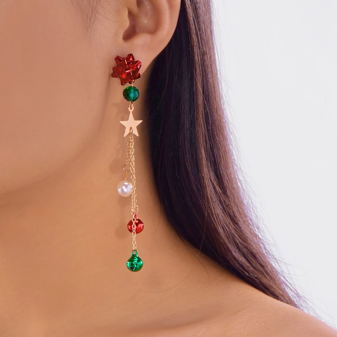 A red bow drop earring with a green bead, gold-tone star, white bead, and dangling bells is worn against dark hair.