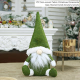 Green fabric Christmas gnome with a pointed hat and white beard sits before a decorated fireplace beside a 16 × 31 cm label.