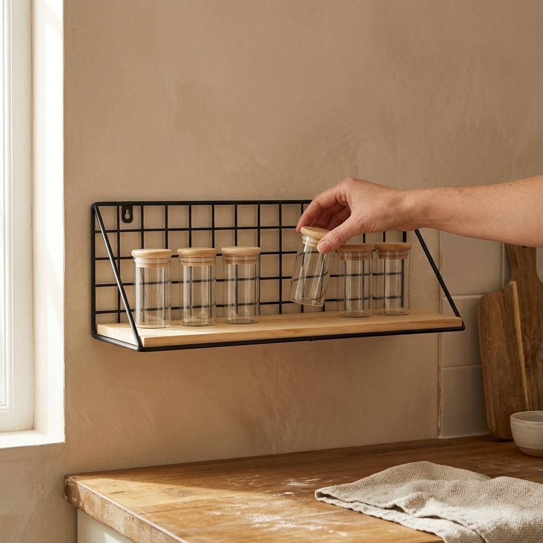 A wall-mounted black wire grid shelf with a light wood base holds several clear glass jars with wooden lids in a kitchen setting. A hand is reaching in to adjust one of the jars.