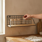 Hand placing a glass spice jar onto the black wire grid wall shelf above a wooden kitchen worktop