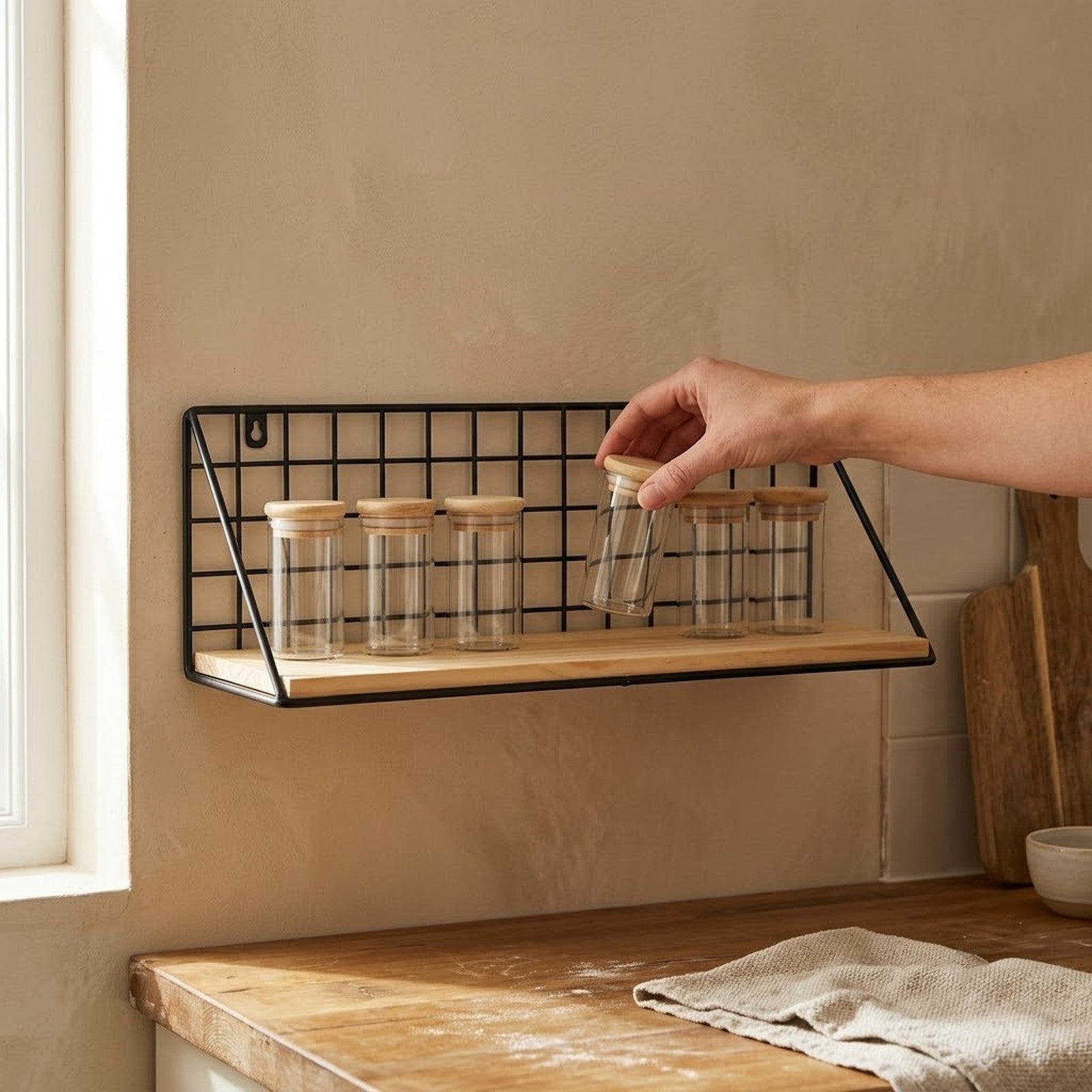 Hand placing a glass spice jar onto the black wire grid wall shelf above a wooden kitchen worktop