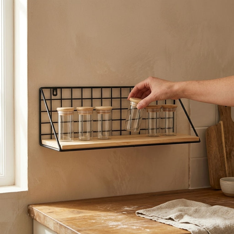 Hand placing a glass spice jar onto the black wire grid wall shelf above a wooden kitchen worktop