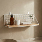 White wall mounted wire shelf in a bathroom holding a soap dispenser, a ceramic jar and a folded towel