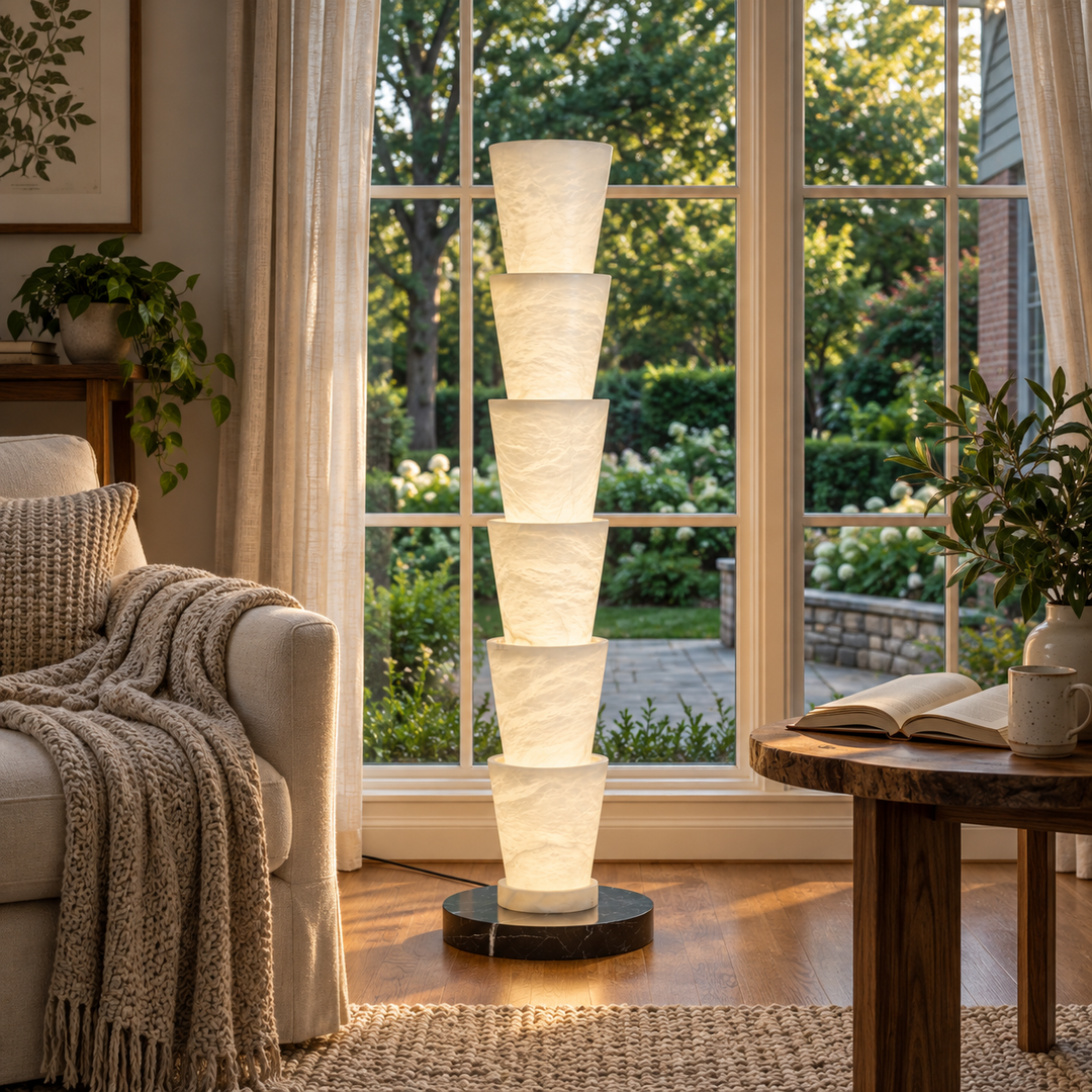 A tall, modern floor lamp with six stacked, tapered white fabric shades on a round black base, standing in a cozy living room next to a window with a garden view.