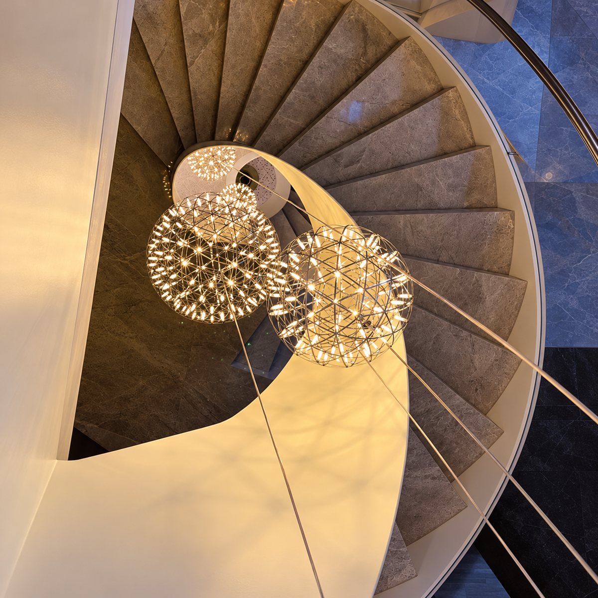 Globe pendant light grouping with illuminated metal lattice frames hangs through the center of a curved staircase.