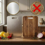 Two rectangular cutting boards with cutout handles are shown on a kitchen counter: one brushed stainless steel and one natural wood, with food items around them. Two circular callout graphics at the top show a mirror on the plain board and a close-up of wood shavings crossed out, suggesting the board is self-cleaning or resistant to staining.