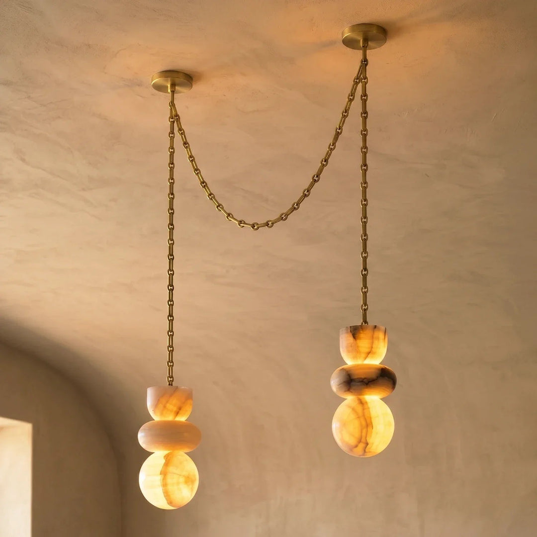 An alabaster chandelier has two glowing, veined stone drops at different heights, joined by a draped gold-toned chain against beige walls.