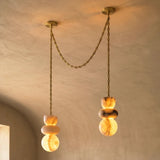 An alabaster chandelier has two glowing, veined stone drops at different heights, joined by a draped gold-toned chain against beige walls.