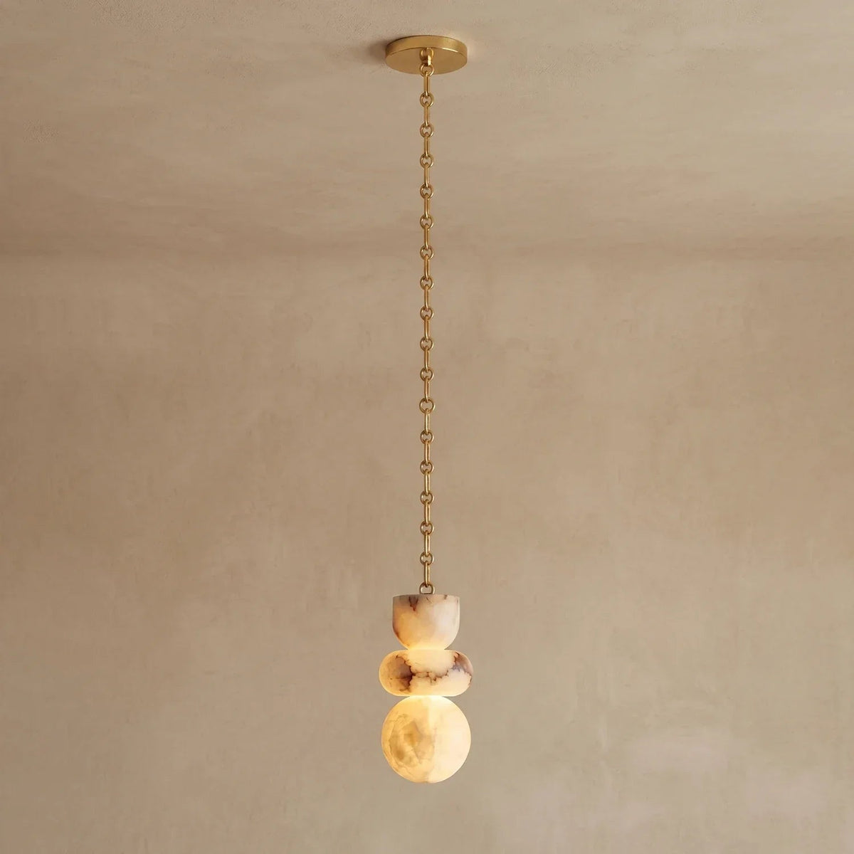 An alabaster chandelier with three stacked cream stone shapes hangs from a gold-toned chain against a beige wall.