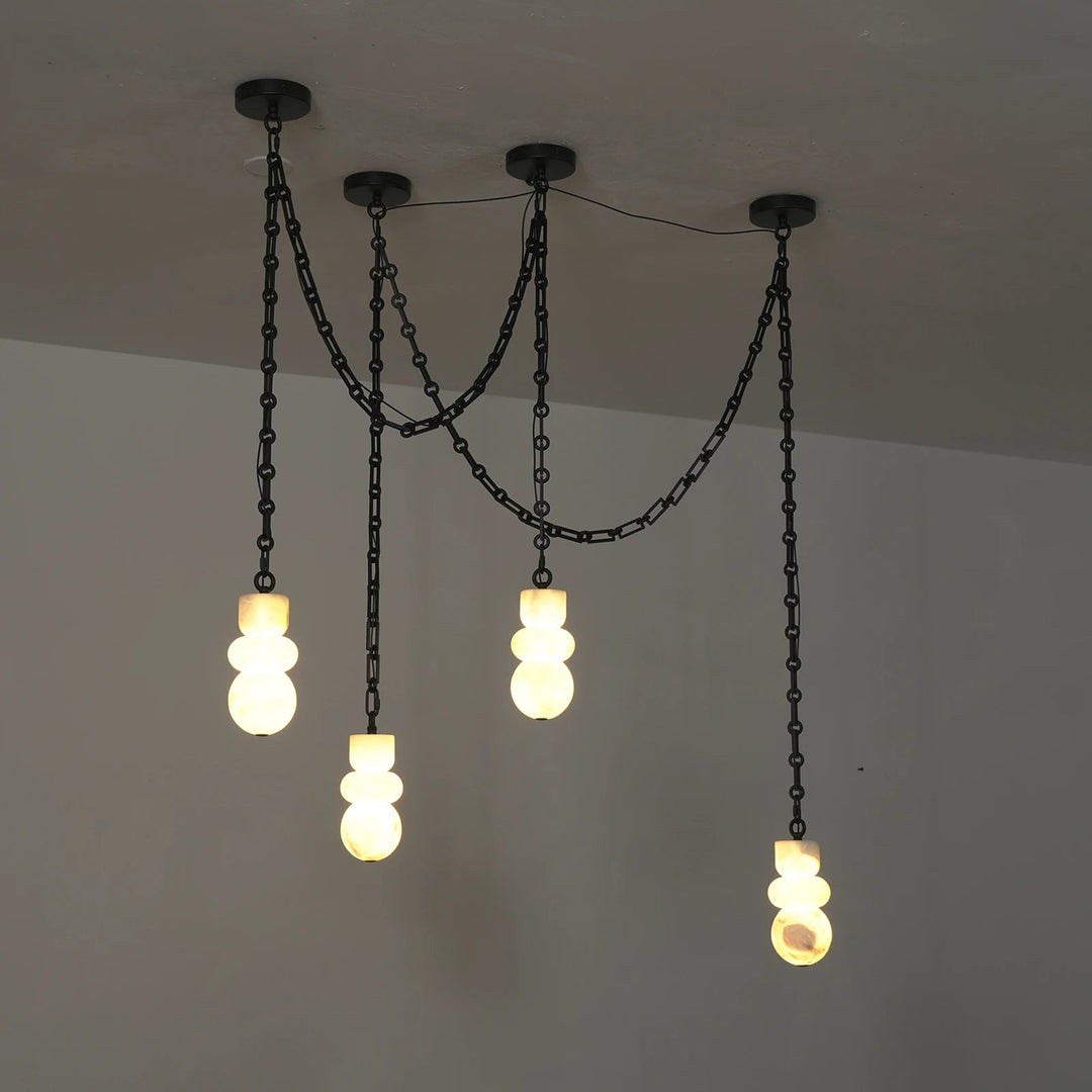 Chandelier with four glowing alabaster shades suspended at staggered heights from draped black chains, viewed at an angle against gray walls.