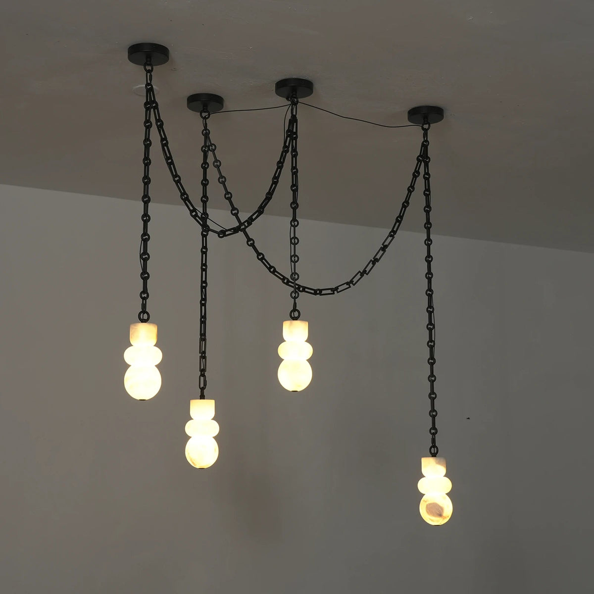 Chandelier with four glowing alabaster shades suspended at staggered heights from draped black chains, viewed at an angle against gray walls.