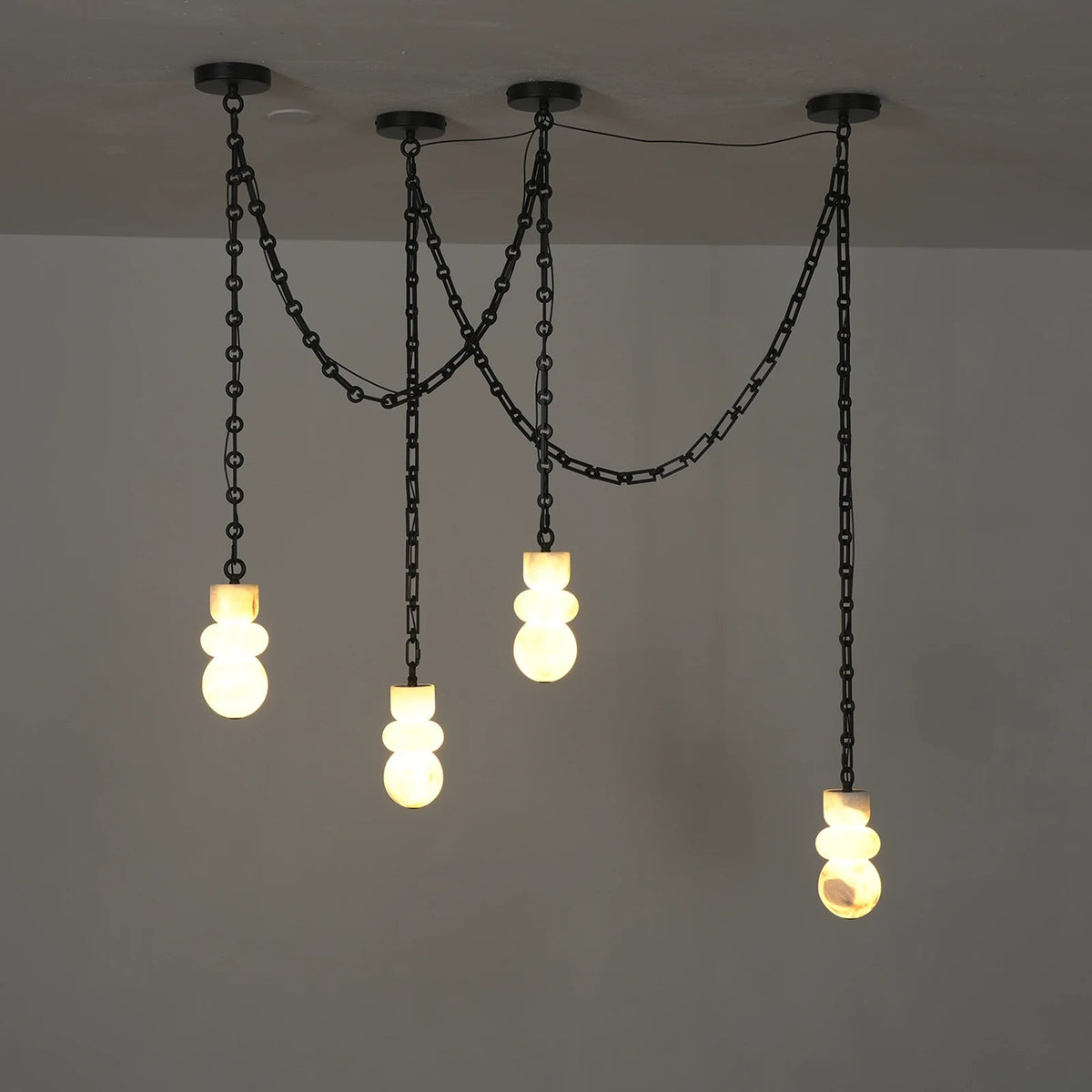 Front view of a chandelier with four illuminated cream alabaster shades and looping black chains against a plain gray wall.