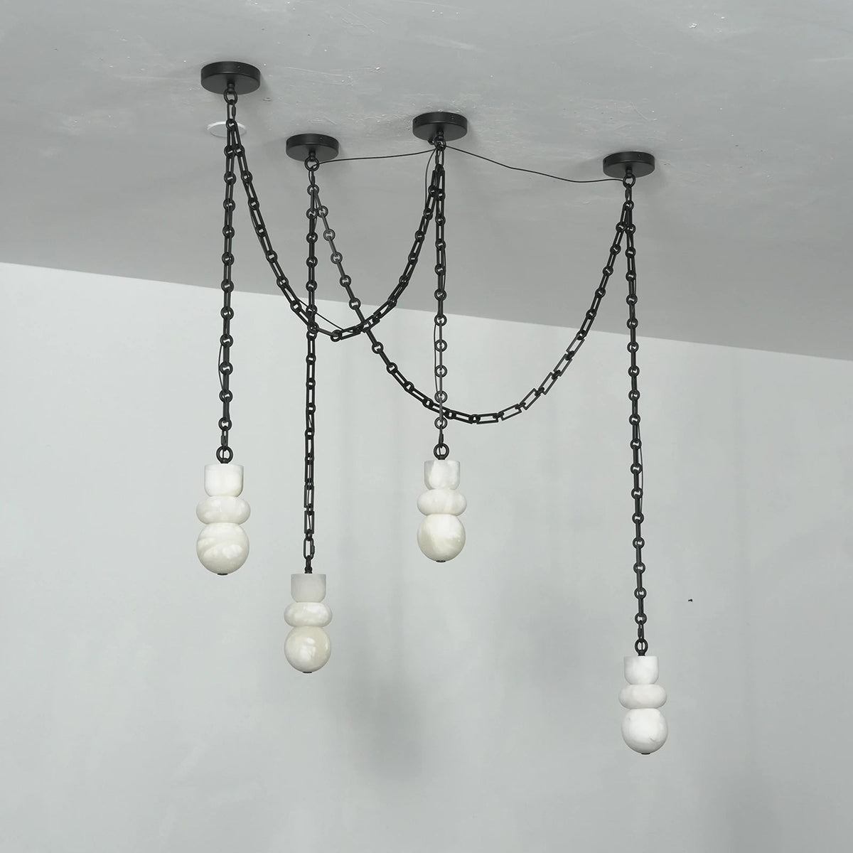 Angled view of an unlit chandelier with four white alabaster shades hanging at different heights from black chains against pale walls.