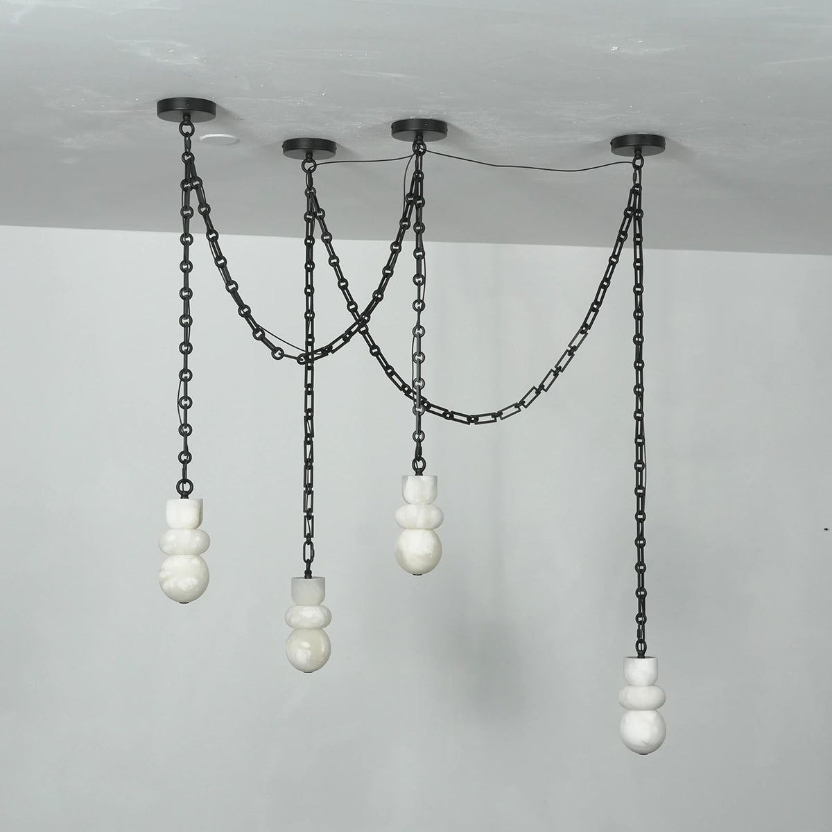 Front view of an unlit chandelier with four sculpted white alabaster shades, draped black chains, and round black ceiling mounts.