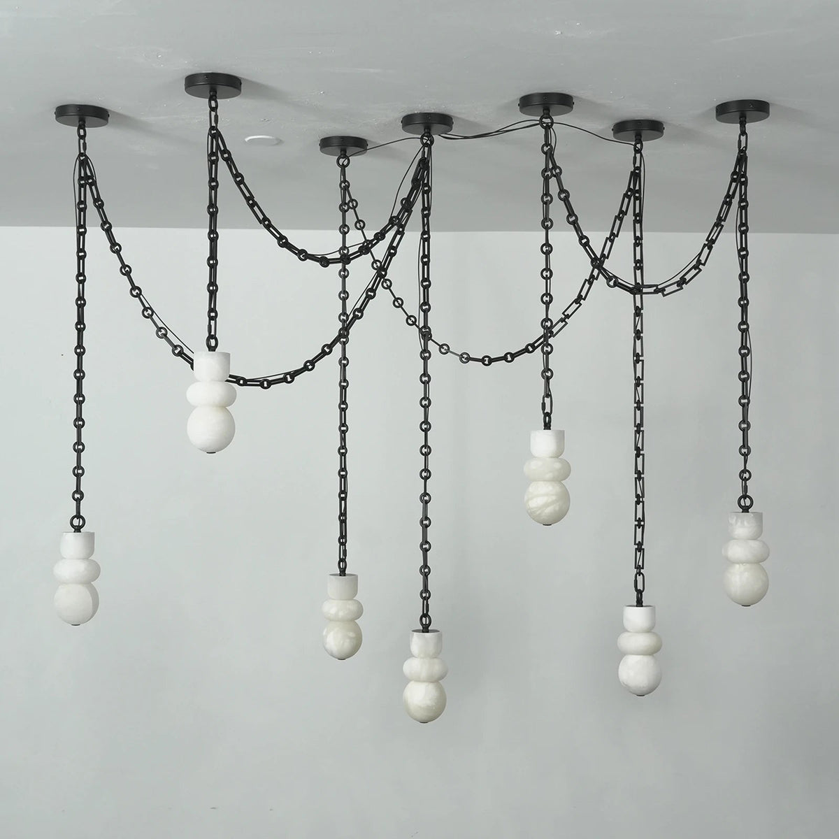 A modern multi-light linear chandelier with black metal chains and spherical white glass shades, suspended from a ceiling against a plain light gray wall.