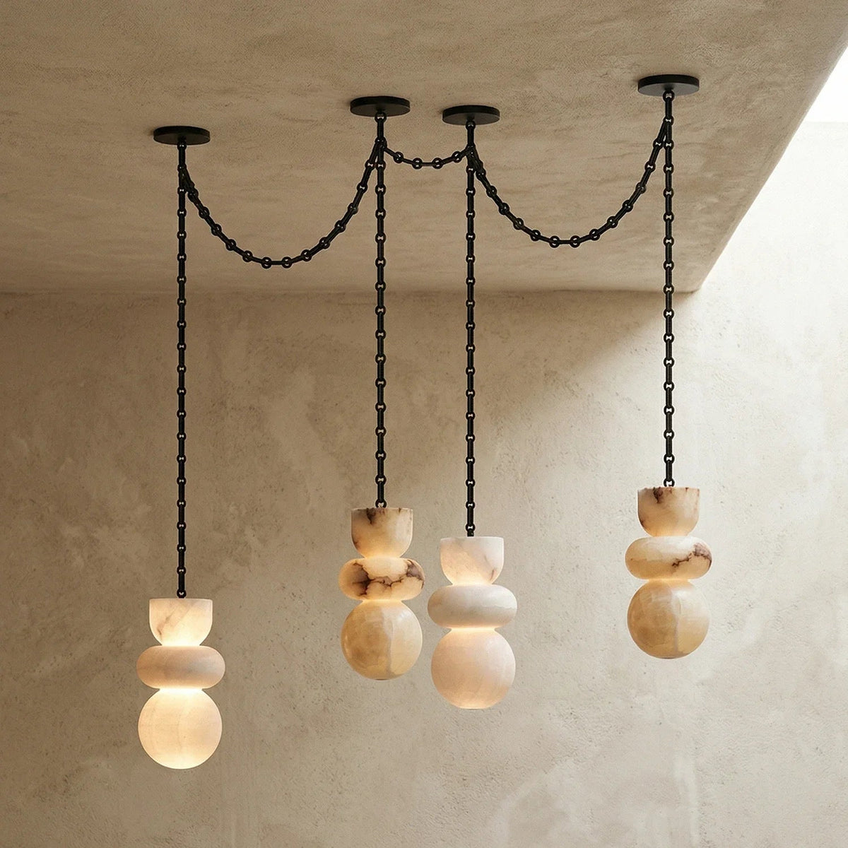 Alabaster chandelier with four glowing stone drops at staggered heights and black chains against textured cream walls.