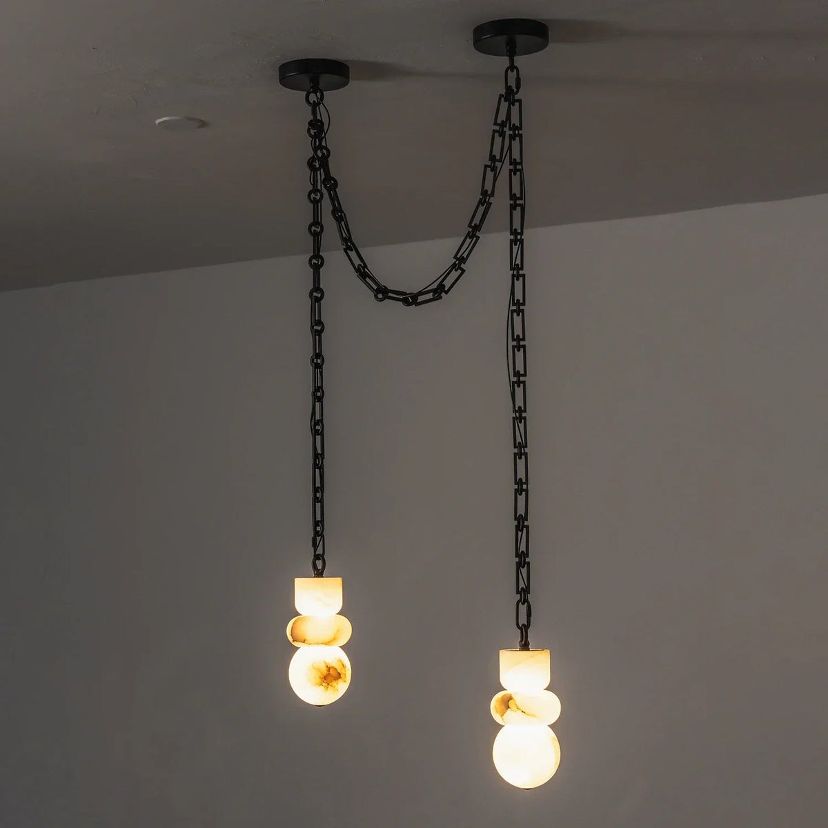 Alabaster chandelier has two glowing drops at different heights, suspended from black chains against a gray wall.