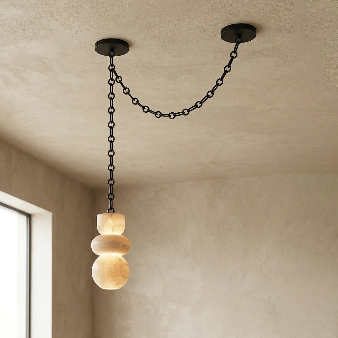 Chandelier with one glowing cream alabaster shade and a black chain swag between two ceiling mounts beside a window.