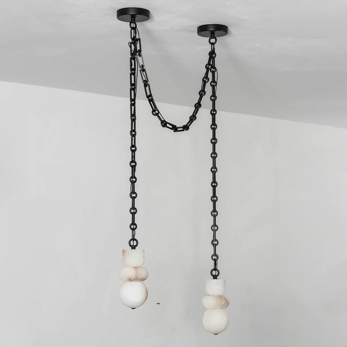 Alabaster chandelier with two white sculpted shades hanging at different heights from black swag chains against a pale wall.