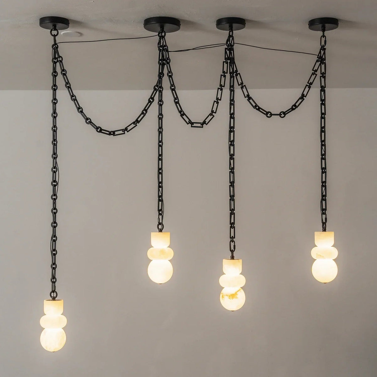 Four-light alabaster chandelier with glowing cream shades and black chains draped between ceiling mounts against a gray wall.
